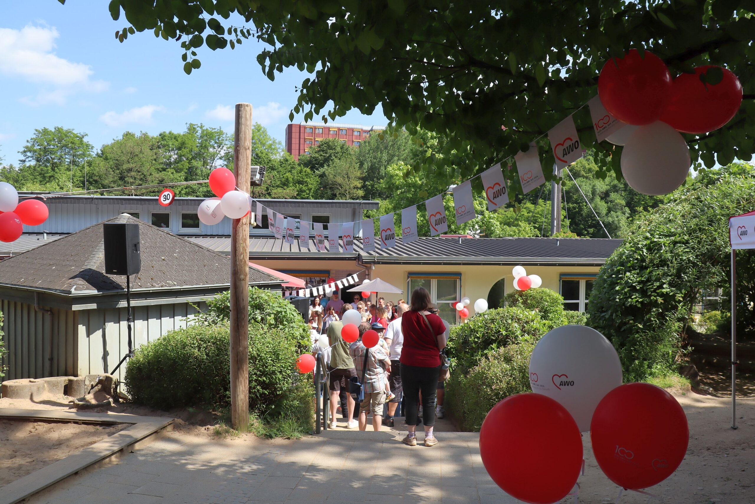 Die Außenanlage der Einrichtung ist mit Luftballons und Fähnchen in den AWO Farben rot und weiß geschmückt. Es sind viele Leute, die die Jubiläumsfeier besuchen, zu sehen.