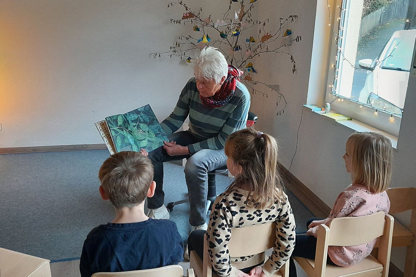 Eine ältere Dame zeigt Kita-Kindern eine Abildung im Bilderbuch. Die Kinder sitzen auf Stühlen im Halbkreis vor ihr.