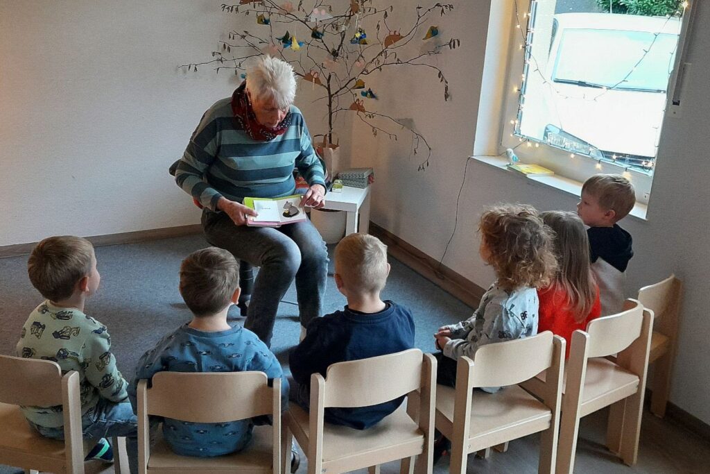 Eine ältere Dame zeigt Kita-Kindern eine Abildung im Bilderbuch. Die Kinder sitzen auf Stühlen im Halbkreis vor ihr.