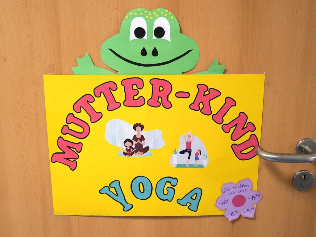 Ein gelbes Schild mit einem großen grünen Frosch aus Papier lädt zum Mutter-Kind-Yoga ein.