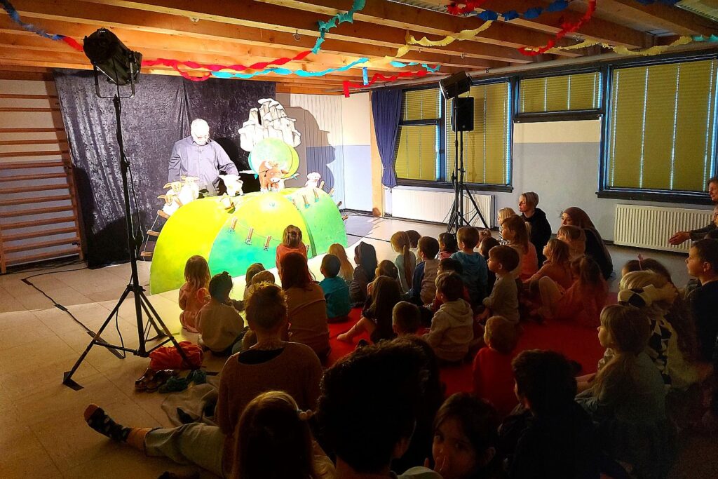 Die Kinder schauen dem Theater zu. Alle KitaKinder sitze auf dem Boden, vorne ist die Bühne aufgebaut Ein Puppenspieler bewegt Stofftiere und erzählt eine Geschichte.