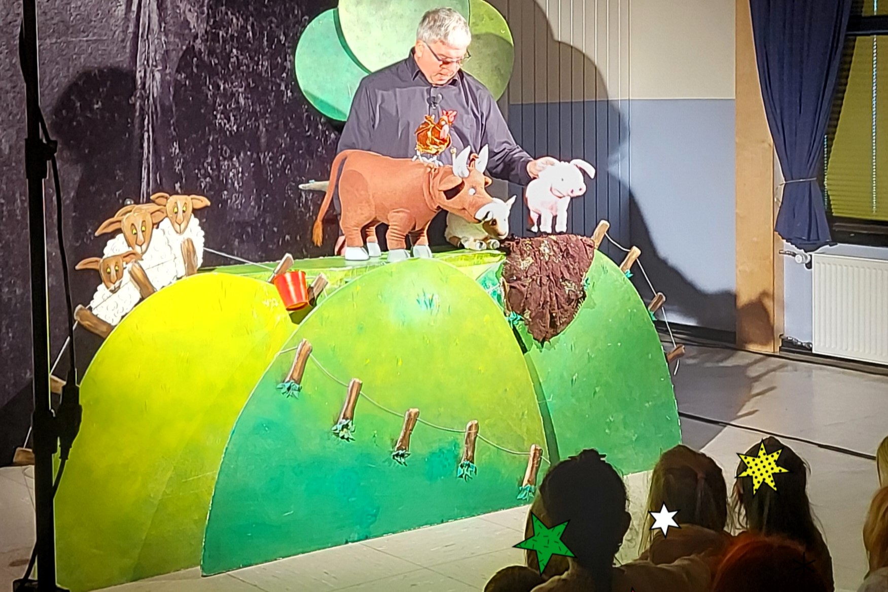 Theater_Käthe Strobel 2025 2 Die Kinder schauen einem Theater zu. Ein Mann spielt eine Kuh, ein Schaf und ein Schwein, die auf einer Wiese stehen.