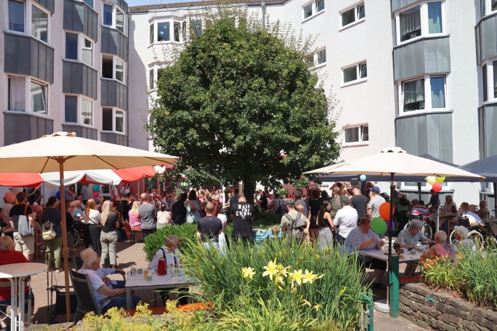 Im Innenhof des AWO Altenzentrums stehen Schirme, Sitzbänk und Tische. Viele Menschen stehen und sitzen dort beim Sommerfest.