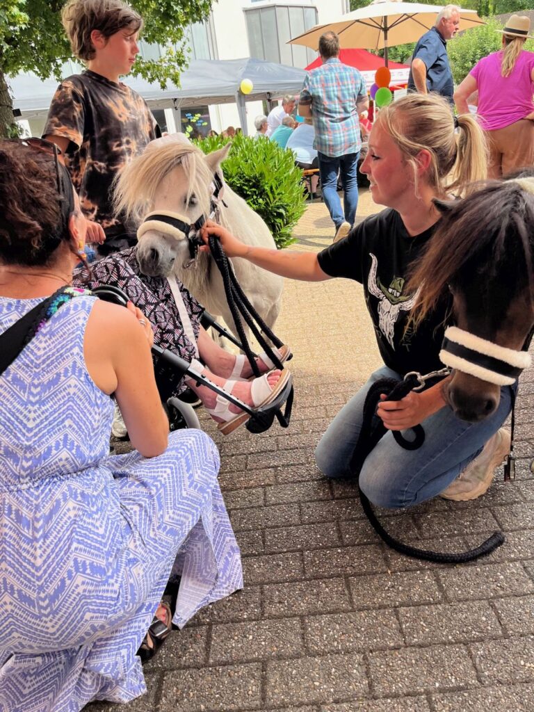 Eine Frau hält ein kleines Pony an der Leine. Sie hockt mit dem Pony vor einer Dame, die im Rollstuhl sitzt.