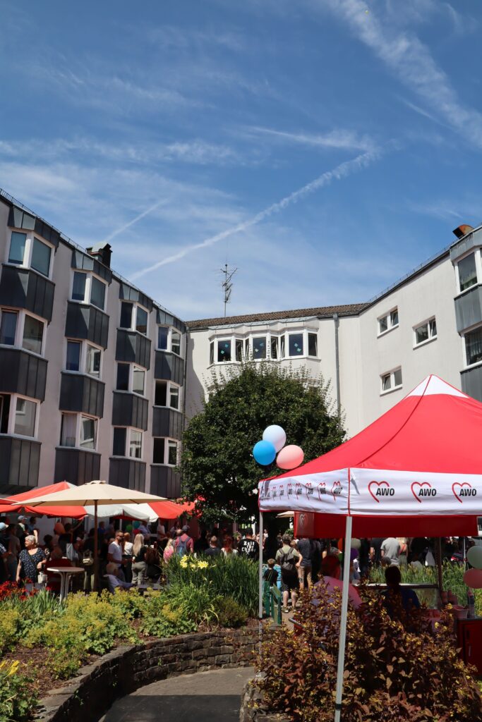 Im Innenhof des AWO Altenzentrums stehen Schirme, Sitzbänk und Tische. Viele Menschen stehen und sitzen dort beim Sommerfest.