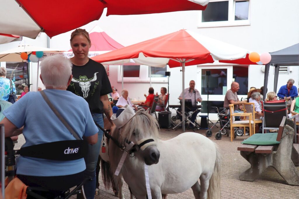 Eine Frau hält ein Zwergpony an der Leine. Das Pony hat einen Matrosenhut auf. Die Frau unterhält sich mit einer Dame, die auf ihrem Rollator sitzt.