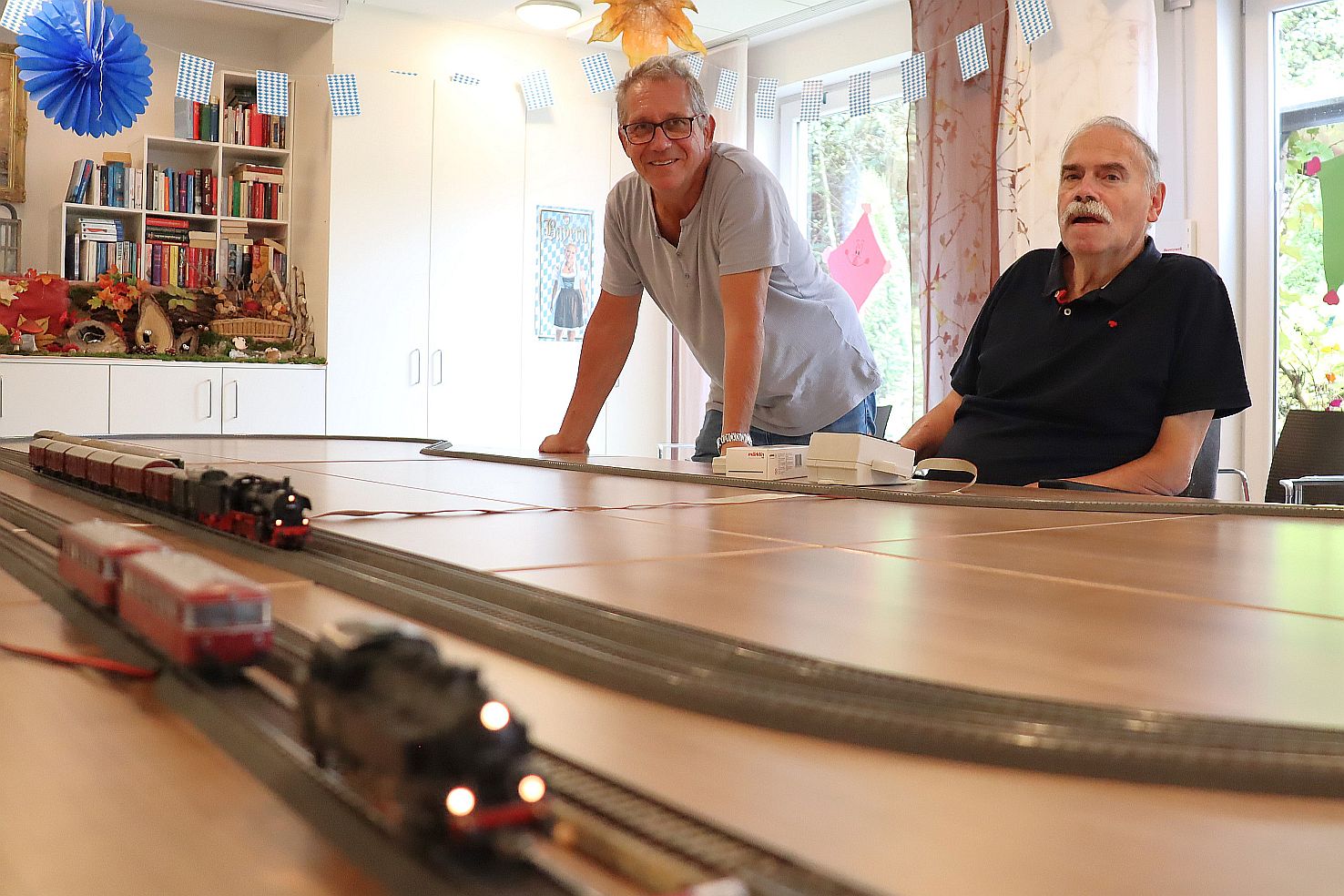Zwei Herren am Tisch lachen in die Kamera. Auf dem Tisch fährt eine Modelleisenbahn auf Schienen
