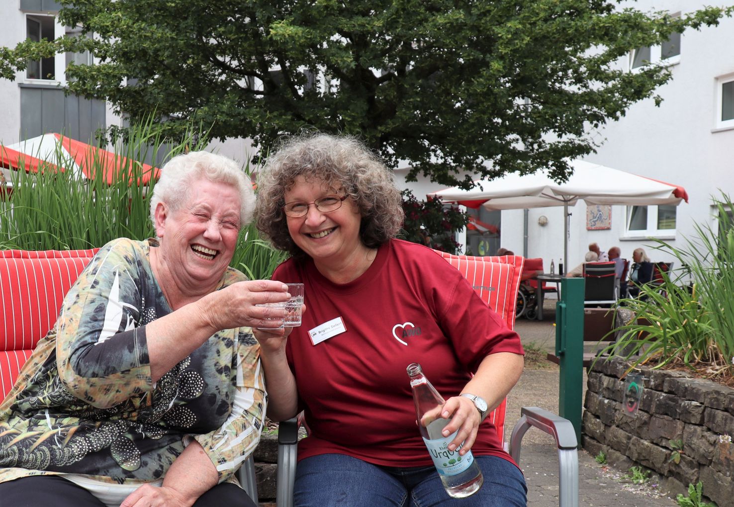 Eine Mitarbeiterin und eine ältere Dame sitzen im Garten nebeneinader. Sie lachen und prosten sich mit Wasser zu